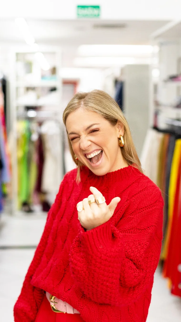 Júlia Wolff sorrindo e piscando no corredor do seu brechó, de suéter vermelho
